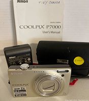 Nikon Coolpix P7000 camera front, showing lens with optical zoom and 14.2MP sticker, charger, black carrying case, and user's manual.