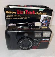 Front view of Nikon Tele Touch Deluxe camera showing lens, flash, and branding with original box in background