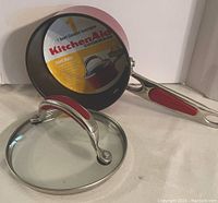 Photo showing KitchenAid 1 Quart Covered Saucepan with glass lid and metal handle with red grip, black interior cooking surface, and the seller's label visible inside the pan.