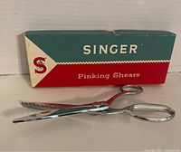 Singer pinking shears displayed in front of their original box, shiny metal with zigzag blades.