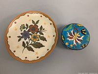 Top-down view of the unsigned pottery pedestal plate and colorful round trinket box side by side on gray surface