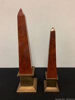 Front view of both obelisk statues side by side showing lacquered wood texture and brass bases
