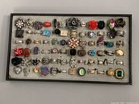Full tray view of over 60 assorted vintage costume rings with floral, geometric, and gemstone designs in multiple colors and metal finishes.