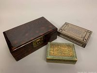 Three decorative boxes shown together on neutral background: dark Chinese lacquer box, Persian marquetry mosaic box with Islamic motif, and smaller Italian Florentine box with gold and green tone wear.