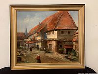 Full view of framed oil painting depicting village street buildings, figures and frame.