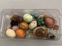 Image showing a clear plastic bin filled with assorted polished stone eggs and figurines including turtle, elephants, birds, and eggs in various colors and patterns.