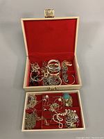 Open jewelry box showing assorted chains, bangles, rings, faux pearl strands, and decorative pendants inside red velvet compartments.