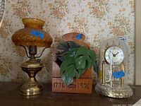 Front view of the three items together: the metal lamp with amber glass globe, the wooden box with faux greens, and the Loricron anniversary clock under dome.