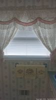 Window with white lacy frilly curtains with pink accents and embroidered towels hanging on a towel rack below the window.