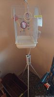 White metal bird cage with toys inside and mounted on a white tripod stand.