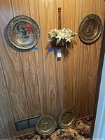 Four brass decorative wall plates mounted on wood panel wall, with a brass wall pocket holding artificial lilies in the center between two plates. Two more plates rest below on the floor.