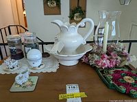 Wide view of lot on wooden surface showing ceramic pitcher and bowl, candle jars, faux flowers, bird figurine, and white egg-shaped ornamental item