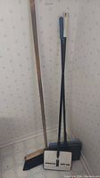 Photo showing two carpet sweepers and a push broom with wooden handle, standing against wall on tiled floor.