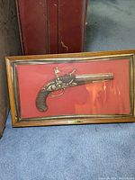 Full view of framed faux flintlock pistol on red background with wood frame and glass cover.