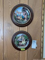 Two Hummel plates vertically mounted on wooden wall, each with a dark wood round frame, upper plate shows two children sitting under a tree and a small rabbit nearby, lower plate shows a boy and girl sitting together outdoors.