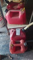 Four red plastic gasoline containers of various sizes arranged on a wooden surface and floor, showing dirt and wear.