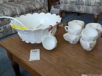 Milk glass punch bowl on a wooden table with 9 cups wrapped in paper and a ladle inside bowl.