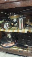 Stainless steel percolator, stacked pots, non-stick skillet, and 12-cup muffin tin visible on a shelf with checkered liner.