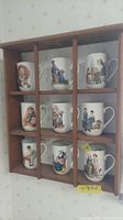 Frontal view of wooden shelf filled with 9 Norman Rockwell ceramic mugs showing varied artwork in each cubby compartment.
