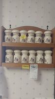 Full view of 12 ceramic spice jars with farm house designs on two-tier wooden rack on wallpapered wall.