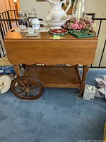 Front view of wooden tea cart showing drop leaf top closed, large wooden wheels, lower shelf, and various items on top (not included).