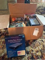 Cardboard box containing assorted VHS tapes and the Analog Pass-Through DTV Converter Box product box placed on carpeted floor.