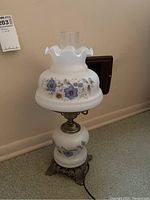 Photo of vintage milk glass hurricane lamp showing front view on carpet, with floral blue rose pattern on glass and ruffled chimney top.
