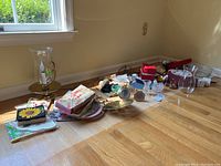 Wide view of the lot contents including brass candle holder with hurricane glass, glass bud vases, plates, napkins, candles, and snuffer spread out on floor near window