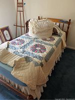 Twin bed dressed with a quilt featuring red and blue floral patchwork, a dust ruffle, and a cream storage bag containing pillows and sheets on top.