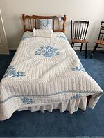 Twin bed made up with white quilt featuring blue floral appliques, white dust ruffle with blue floral prints, white pillow, and sheets.