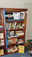Front view of the tall wood bookcase filled with books and boxed items to show shelving and condition.