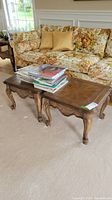 Pair of wooden end tables with carved accents placed in front of a floral upholstered couch on a beige carpet. One table has magazines stacked on top.
