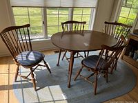 View of the dining table with four wooden spindle-back chairs placed on an oval light blue rug near large windows. Chairs have striped blue cushions.
