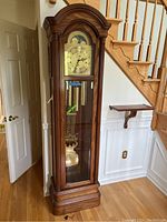 Full view of Heritage Heirlooms grandfather clock with wooden case and glass door, showing pendulum and weights inside.