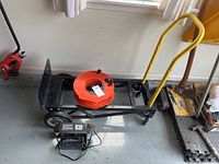 Folding hand truck with yellow handle, black frame, and two wheels, with an orange cord reel extension cord placed on it and Schumacher 1 amp electric battery charger on floor.
