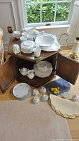 Full view of ironstone china set displayed on a wooden hexagonal cabinet and the floor around it with white teacups, saucers stacked on top, octagonal serving bowls, plates, gravy boat with under plate, creamer, sugar bowl, and salt and pepper shakers all visible.