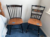 Two Hitchcock style chairs side by side, one with floral backrest and one with spindle backrest, both with natural wood seats and black painted legs and spindles