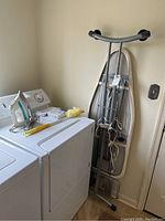 Iron, ironing board folded and standing against wall, iron and extension cord on dryer top, with cleaning supplies.
