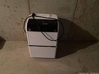 White Kenmore dehumidifier with front vent and black top panel, power cord wrapped on top, photographed in a basement setting.