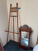 Full view of the wood easel and wood-framed wall mirror leaning against the wall on carpeted floor.