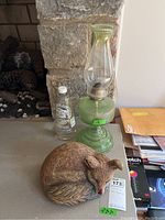 Green glass oil lamp with clear chimney and metal burner next to a bottle of lamp oil and small curled resin fox statuette on a stone fireplace hearth.