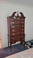 Front view of a tall wood highboy dresser with curved bonnet top and brass handles showcasing five drawers on the upper section and three on the lower section.