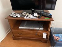Front view showing wooden stand with one dovetail drawer and open shelf, visible scratches, and items on top including multiple remote controls and a TV above.