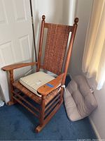 Full view of wooden rocking chair with wicker seat and backrest. Attached beige seat cushion with ties and loose beige cushion alongside the chair.