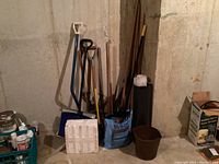 Photo showing collection of garden tools including shovels, pitchforks, stamper, bucket, and bags of solar salt and plastic containers against basement wall.