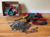 Overview of the vintage Lionel train set including the locomotive, several train cars in red, green, and yellow, a box with green Christmas-themed items, and a large pile of curved and straight metal track pieces on the floor
