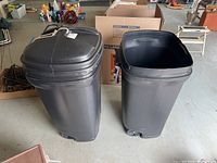 View of two black plastic trash cans, one with lid on and one open without lid.