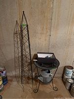 Full view of the metal trellis, birdbath on metal base with items on top, and watering can with paint cans nearby
