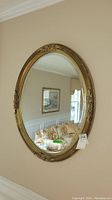 Frontal photo of the ornate oval wall mirror showing the detailed gold-tone carved frame and the beveled glass reflecting a floral sofa and artwork.
