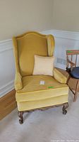 Front view of yellow vintage wingback chair with a beige throw pillow on the seat, showing fabric texture, cushion, and carved wooden legs.